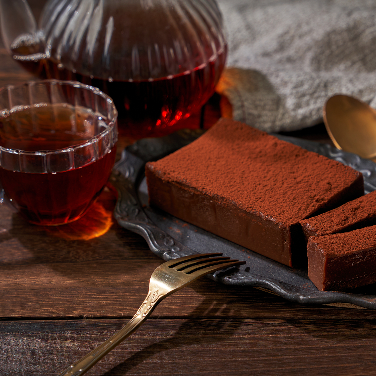 A glass plate holds rectangular slices of Small Batch Magic Valentine’s 2026 chocolate cake dusted with cocoa, beside a vintage fork, chocolate cake tea, and a glass pitcher on a wooden table with a gray cloth background.