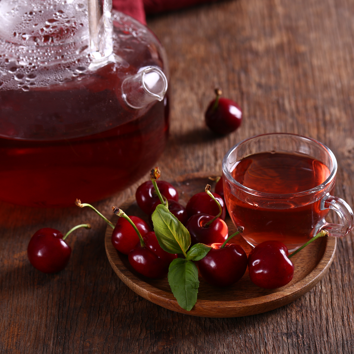 A glass teapot and a glass cup filled with Small Batch Magic Valentine’s 2026 vanilla rooibos cherry tea sit on a wooden table, with fresh cherries and green leaves arranged on a small wooden plate beside the cup.
