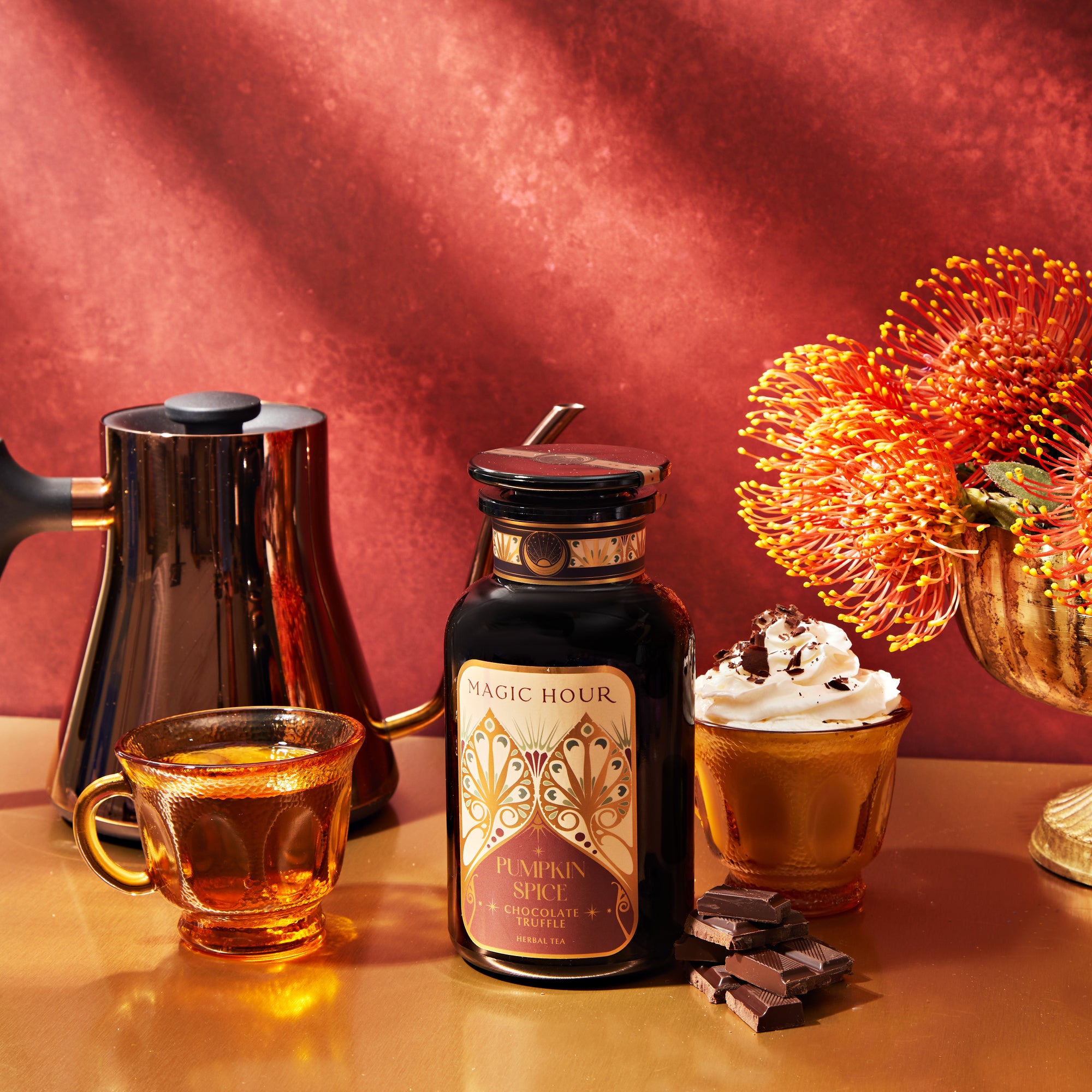 An inviting autumn-themed arrangement with the Pumpkin Spice Essentials Tea Trio Gift Set, showcasing pumpkin spice chocolate tea, a stylish kettle, a clear mug filled with a whipped cream-topped hot drink, pieces of dark chocolate, and bright orange flowers on a warm-toned table against a deep red background.