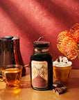 An inviting autumn-themed arrangement with the Pumpkin Spice Essentials Tea Trio Gift Set, showcasing pumpkin spice chocolate tea, a stylish kettle, a clear mug filled with a whipped cream-topped hot drink, pieces of dark chocolate, and bright orange flowers on a warm-toned table against a deep red background.