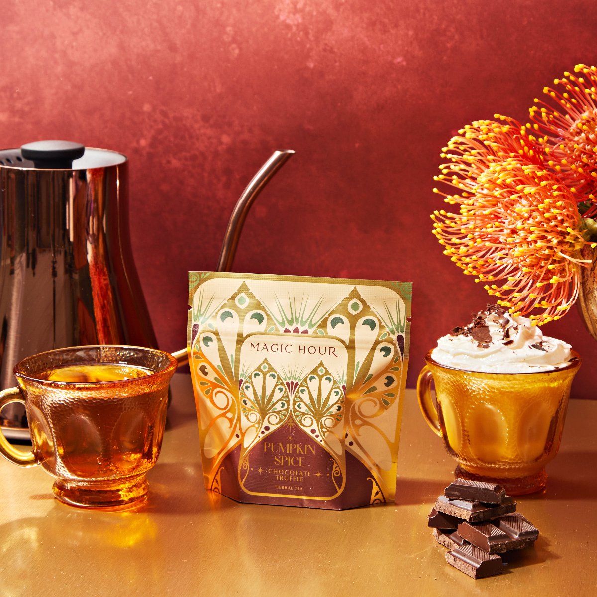 On a table, two amber glass cups sit alongside a decorative package labeled "The Best of Fall Box." One cup holds a pumpkin spice tea while the other features an Apple Crisp beverage topped with whipped cream. Accompanied by a silver teapot and pieces of chocolate, the vibrant orange flower completes the picturesque scene that captures the essence of The Best of Fall Box.