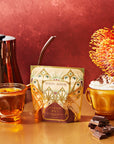 On a table, two amber glass cups sit alongside a decorative package labeled "The Best of Fall Box." One cup holds a pumpkin spice tea while the other features an Apple Crisp beverage topped with whipped cream. Accompanied by a silver teapot and pieces of chocolate, the vibrant orange flower completes the picturesque scene that captures the essence of The Best of Fall Box.