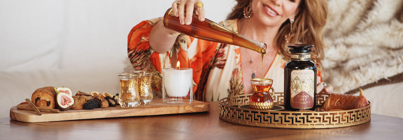 A woman with wavy hair is pouring liquid from a bottle into a glass. She is seated at a table with an assortment of food, including croissants, figs, and honey jars on wooden and decorative trays. The background includes a plush couch and a cozy, fur blanket.