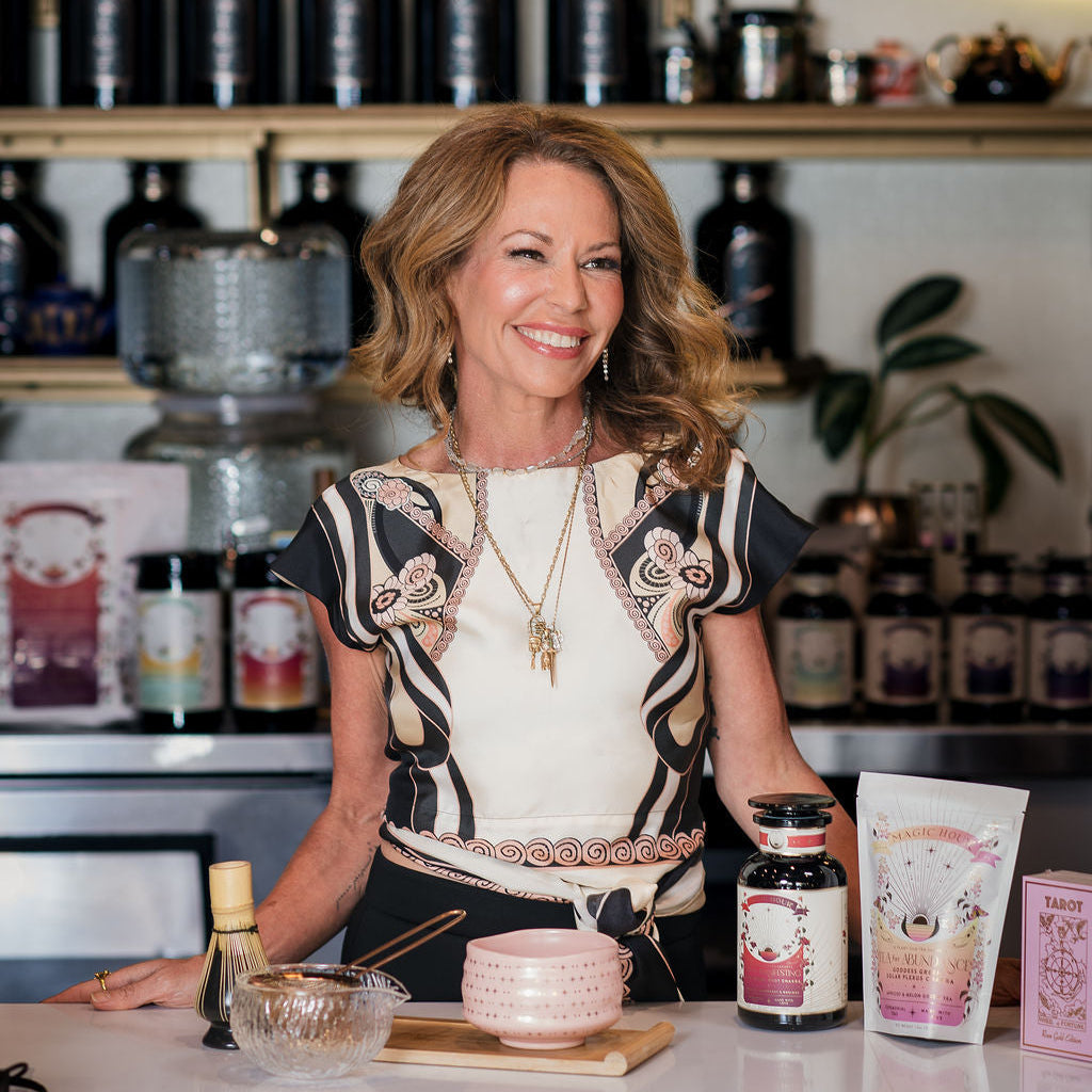 A smiling woman stands behind a counter adorned with beauty products, a bowl, and a whisk—evoking the inviting atmosphere of The Soul of Tea: A Three Day Retreat in Ojai, CA. Shelves lined with bottles and a glowing circular sign create an ambiance that seamlessly blends the sophistication of skincare with the serene rituals celebrated during this immersive Ojai tea retreat.