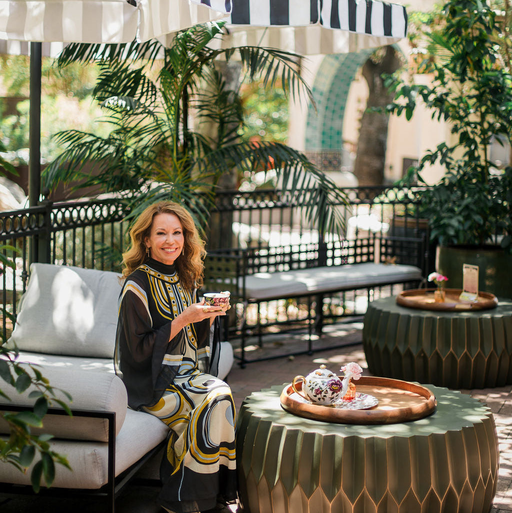 During The Soul of Tea: A Three Day Retreat in Ojai, CA, a woman in a patterned dress sits on an outdoor sofa, smiling and holding a drink amid lush greenery and striped umbrellas at the Ojai Tea Salon. An elegant tray of food and drinks rests on a nearby table as she enjoys the relaxing tea retreat experience.