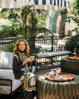 During The Soul of Tea: A Three Day Retreat in Ojai, CA, a woman in a patterned dress sits on an outdoor sofa, smiling and holding a drink amid lush greenery and striped umbrellas at the Ojai Tea Salon. An elegant tray of food and drinks rests on a nearby table as she enjoys the relaxing tea retreat experience.