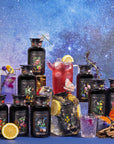 A vibrant display features the Cosmic Garden Iced Tea Sampler Set’s ornate, botanical-labeled bottles, surrounded by glasses of iced tea, citrus slices, flowers, and butterflies, set against a starry galaxy backdrop.