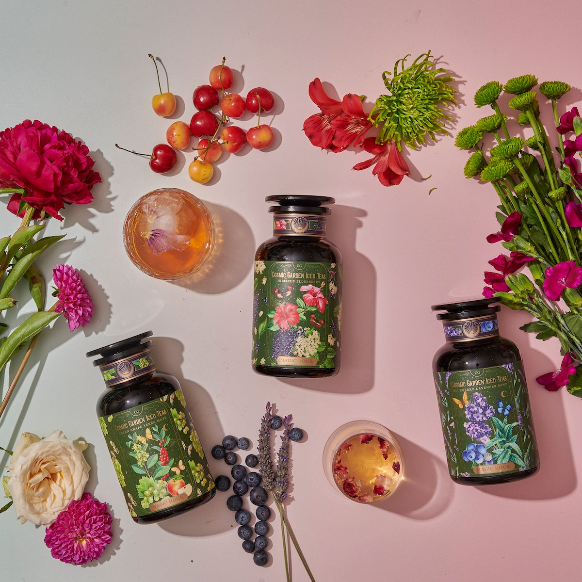 Three bottles of tea with botanical labels, part of the Cosmic Garden Cold Steep Trio Set by Magic Hour, are arranged on a light surface, surrounded by vibrant flowers and fresh fruits like cherries, blueberries, and citrus slices. Two glasses of iced tea are also present, enhancing the colorful and refreshing scene.