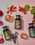 Three bottles of tea with botanical labels, part of the Cosmic Garden Cold Steep Trio Set by Magic Hour, are arranged on a light surface, surrounded by vibrant flowers and fresh fruits like cherries, blueberries, and citrus slices. Two glasses of iced tea are also present, enhancing the colorful and refreshing scene.