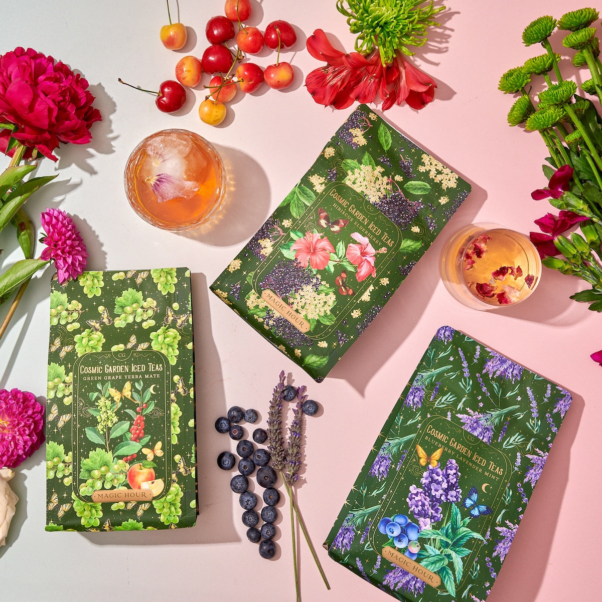 A flatlay photo of three boxes of organic garden herb teas, including the Cosmic Garden Cold Steep Trio Set by Magic Hour, surrounded by fresh flowers, cherries, and blueberries. Two drinks with flower garnishes are placed near the boxes, all set on a pastel-colored surface. The tea boxes feature floral designs.