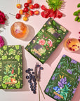 A flatlay photo of three boxes of organic garden herb teas, including the Cosmic Garden Cold Steep Trio Set by Magic Hour, surrounded by fresh flowers, cherries, and blueberries. Two drinks with flower garnishes are placed near the boxes, all set on a pastel-colored surface. The tea boxes feature floral designs.