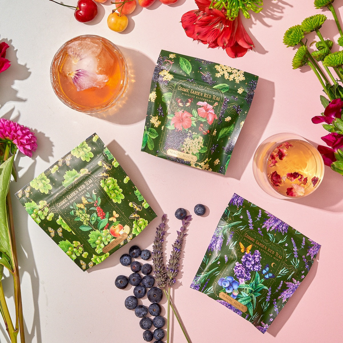 Three vibrant floral tea packages, part of the Cosmic Garden Cold Steep Trio Set by Magic Hour, are surrounded by various flowers, blueberries, and two glass cups filled with drinks. The scene is well-lit, focusing on the colorful and nature-inspired tea packaging.