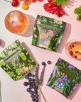 Three vibrant floral tea packages, part of the Cosmic Garden Cold Steep Trio Set by Magic Hour, are surrounded by various flowers, blueberries, and two glass cups filled with drinks. The scene is well-lit, focusing on the colorful and nature-inspired tea packaging.