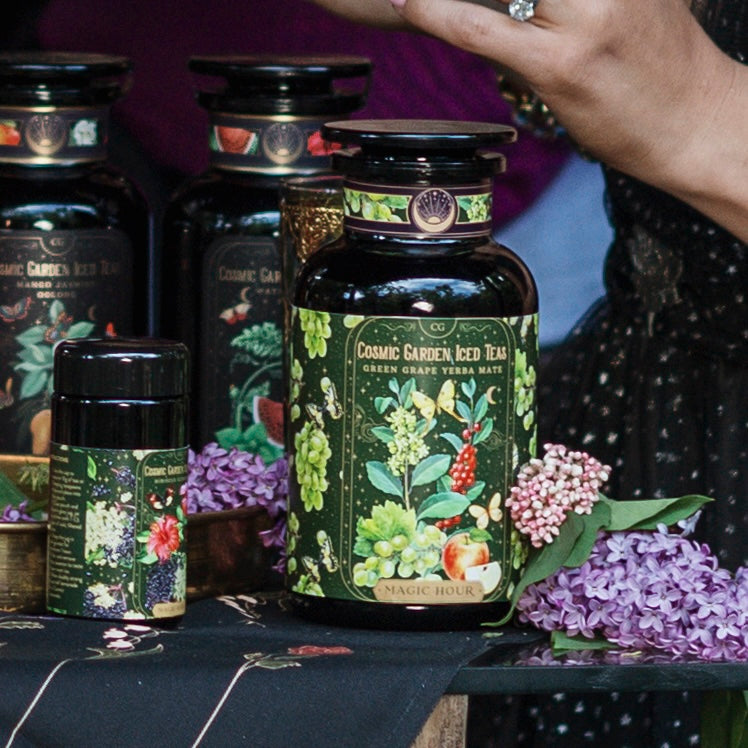Close-up of various Magic Hour tea products adorned with vibrant floral and fruit illustrations. The focal point is a large jar labeled &quot;Green Grape Yerba Mate: Cosmic Garden Iced Tea.&quot; Nearby are fresh lilac blooms, a delicately manicured hand, and an elegant tin of organic loose leaf tea.