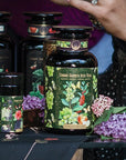 Close-up of various Magic Hour tea products adorned with vibrant floral and fruit illustrations. The focal point is a large jar labeled "Green Grape Yerba Mate: Cosmic Garden Iced Tea." Nearby are fresh lilac blooms, a delicately manicured hand, and an elegant tin of organic loose leaf tea.