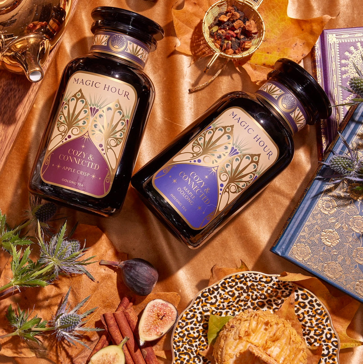 A cozy, autumn-themed flat lay featuring the Cozy &amp; Connected Bundle with two bottles of Autumn teas from &quot;Magic Hour.&quot; One bottle sports a purple label, and the other has a blue label. The scene is adorned with leaves, figs, thistles, a pastry, a teapot, cinnamon sticks, dried tea leaves, and a leopard print plate next to the Cream Ceremonial Tea Bowl.