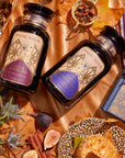 A cozy, autumn-themed flat lay featuring the Cozy & Connected Bundle with two bottles of Autumn teas from "Magic Hour." One bottle sports a purple label, and the other has a blue label. The scene is adorned with leaves, figs, thistles, a pastry, a teapot, cinnamon sticks, dried tea leaves, and a leopard print plate next to the Cream Ceremonial Tea Bowl.