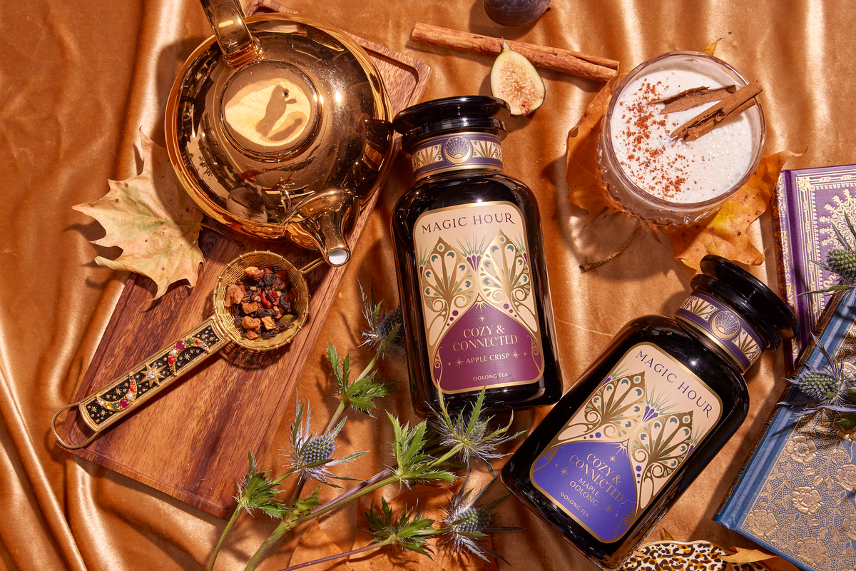 A warm-toned setup featuring a gold teapot, two jars of "Magic Hour" tea, a cup of milk tea with cinnamon sticks, a spoon with tea leaves, figs, a dried leaf, a purple book, and thistle flowers on a gold satin fabric background.