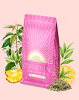 A pink bag labeled "Cream Soda White Tea: Ceremonial Soda Tea" rests amid green leaves, pineapple slices, and loose tea leaves on a pale pink background, evoking the refreshing taste of a sugar-free cream soda white tea blend.