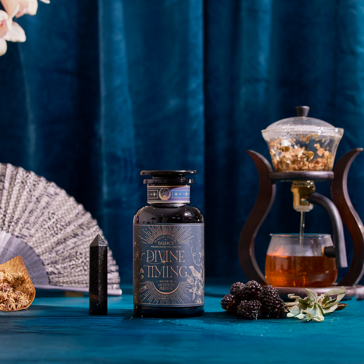 A bottle of Divine Timing Balance rests on a blue table alongside a crystal, dried fruit, blackberries, a fan, and a teapot with tea, set before a luxurious blue velvet curtain.