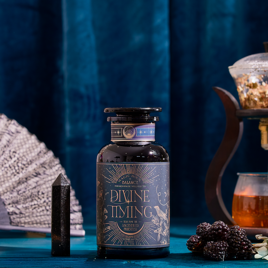 A bottle of Divine Timing Balance rests on a blue table alongside a crystal, dried fruit, blackberries, a fan, and a teapot with tea, set before a luxurious blue velvet curtain.