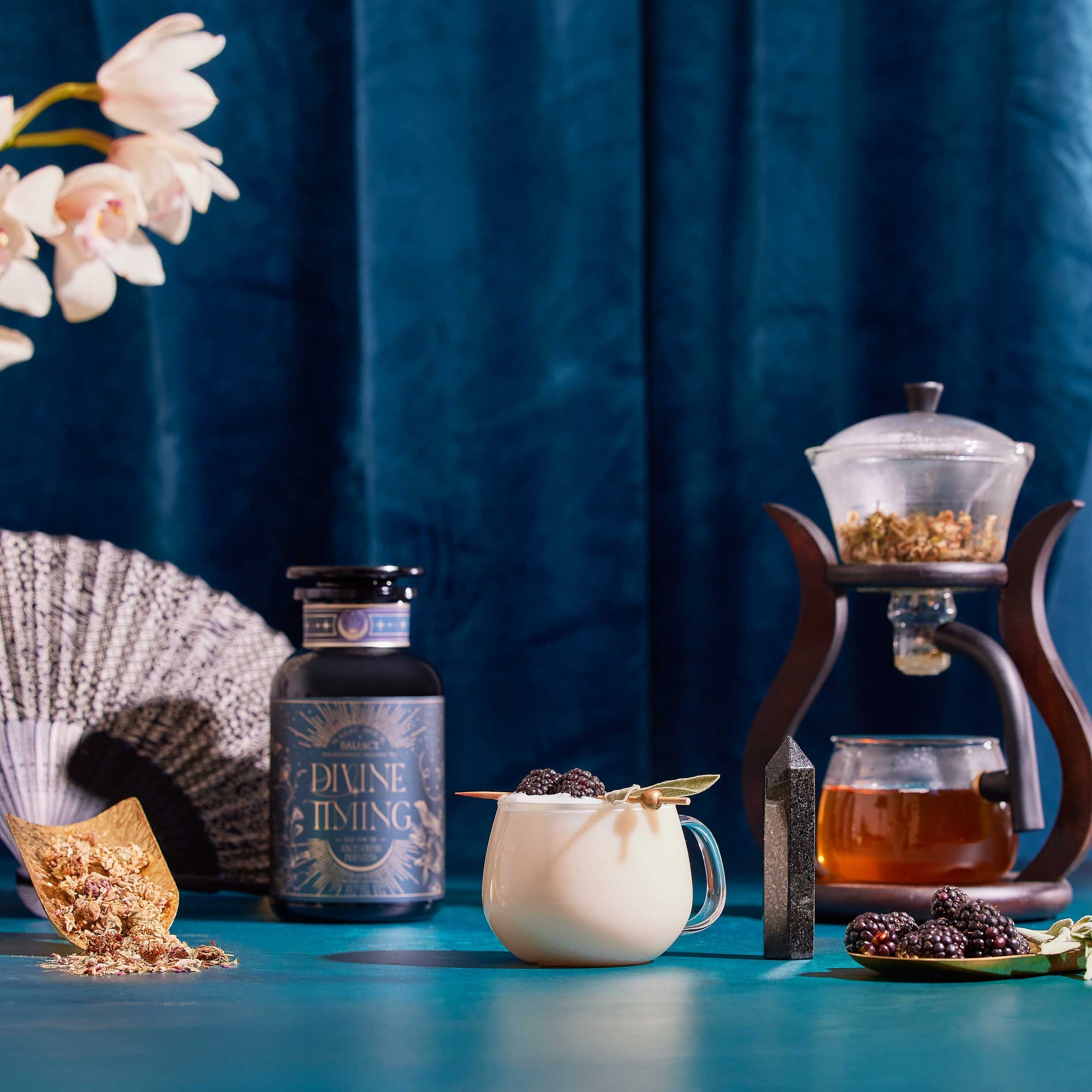 A decorative table displays a white mug with a gold spoon and blackberries beside Divine Timing Balance for menopause support, along with a fan, flowers, gemstones, cooling and dried herbs, all set against a blue curtain.