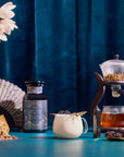A decorative table displays a white mug with a gold spoon and blackberries beside Divine Timing Balance for menopause support, along with a fan, flowers, gemstones, cooling and dried herbs, all set against a blue curtain.