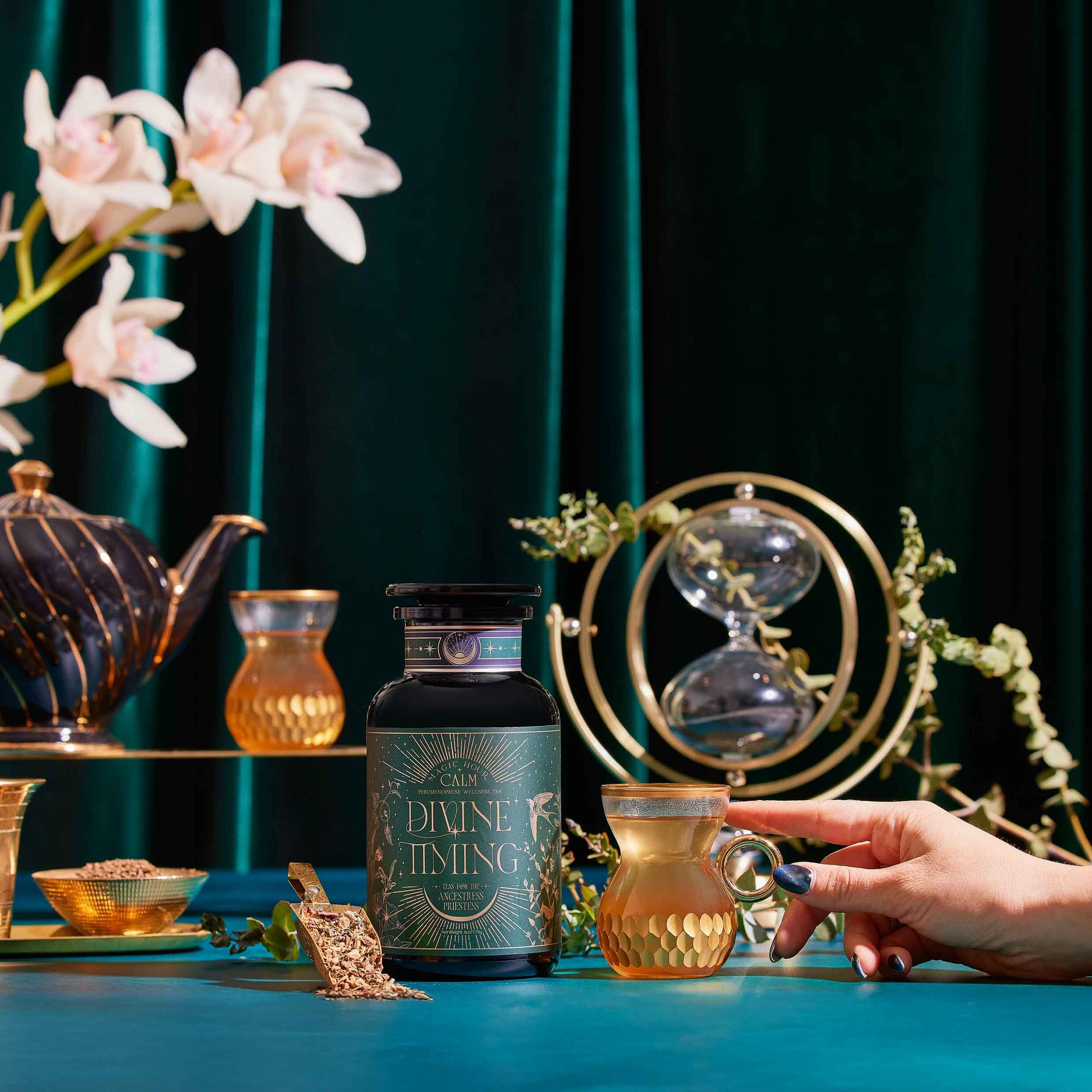 A hand holds a glass of Divine Timing Calm herbal tea near its jar on a teal table with gold accents, alongside a teapot, small cup, hourglass, and white flowers in the background.
