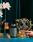 A hand holds a glass of Divine Timing Calm herbal tea near its jar on a teal table with gold accents, alongside a teapot, small cup, hourglass, and white flowers in the background.