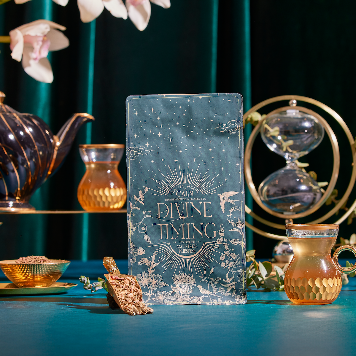 The Divine Timing Calm plaque, featuring gold designs and the words “Calm, Divine Timing, Rest & Trust, Answers and Prayers,” stands on a teal table with teacups, a teapot, an hourglass, and flowers against a green background.