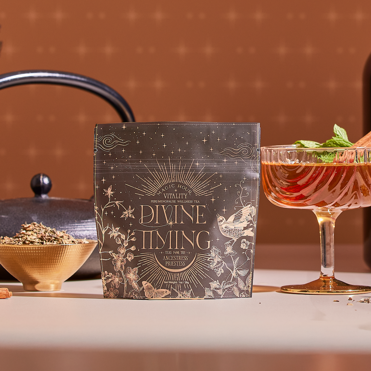 A packet labeled “Divine Timing Vitality” herbal tea sits on a table with loose herbs, a black teapot, and a glass of mint-garnished tea—showcasing its adaptogenic and metabolism-supporting herbs in warm light.