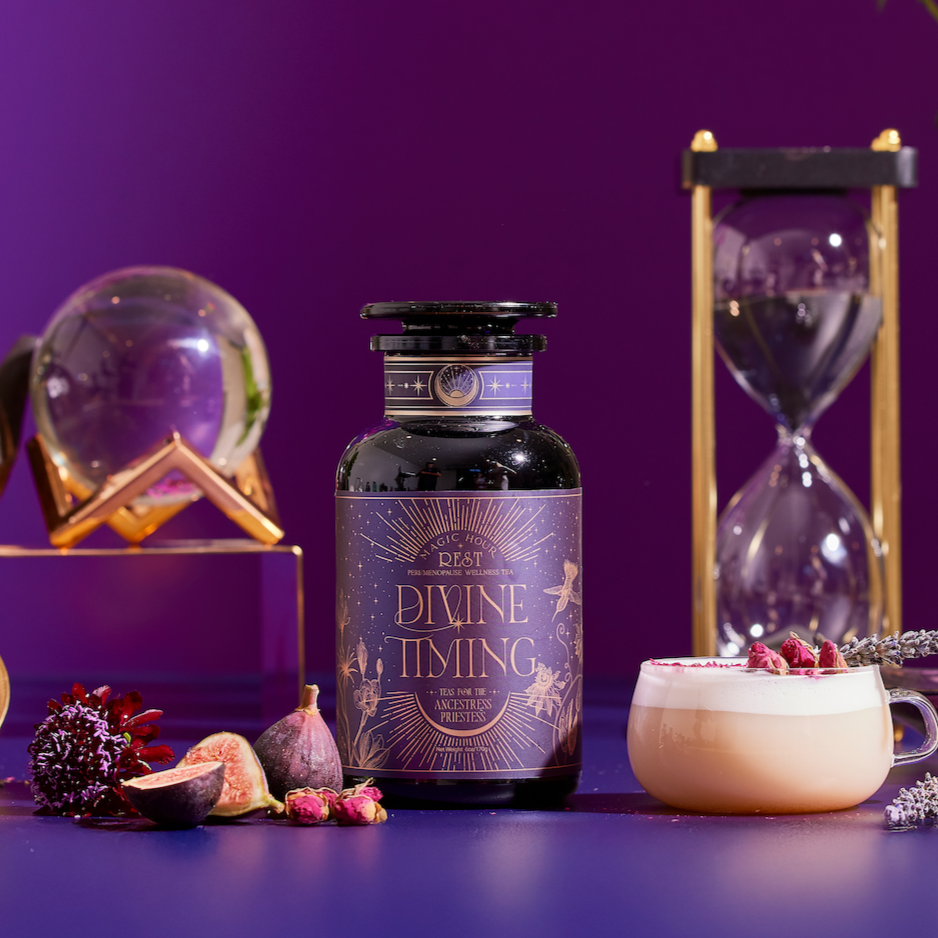 A bottle labeled “Divine Timing Rest” rests on a purple surface with figs, flowers, a latte, hourglass, and glass orb nearby. The purple gradient background adds a mystical atmosphere.