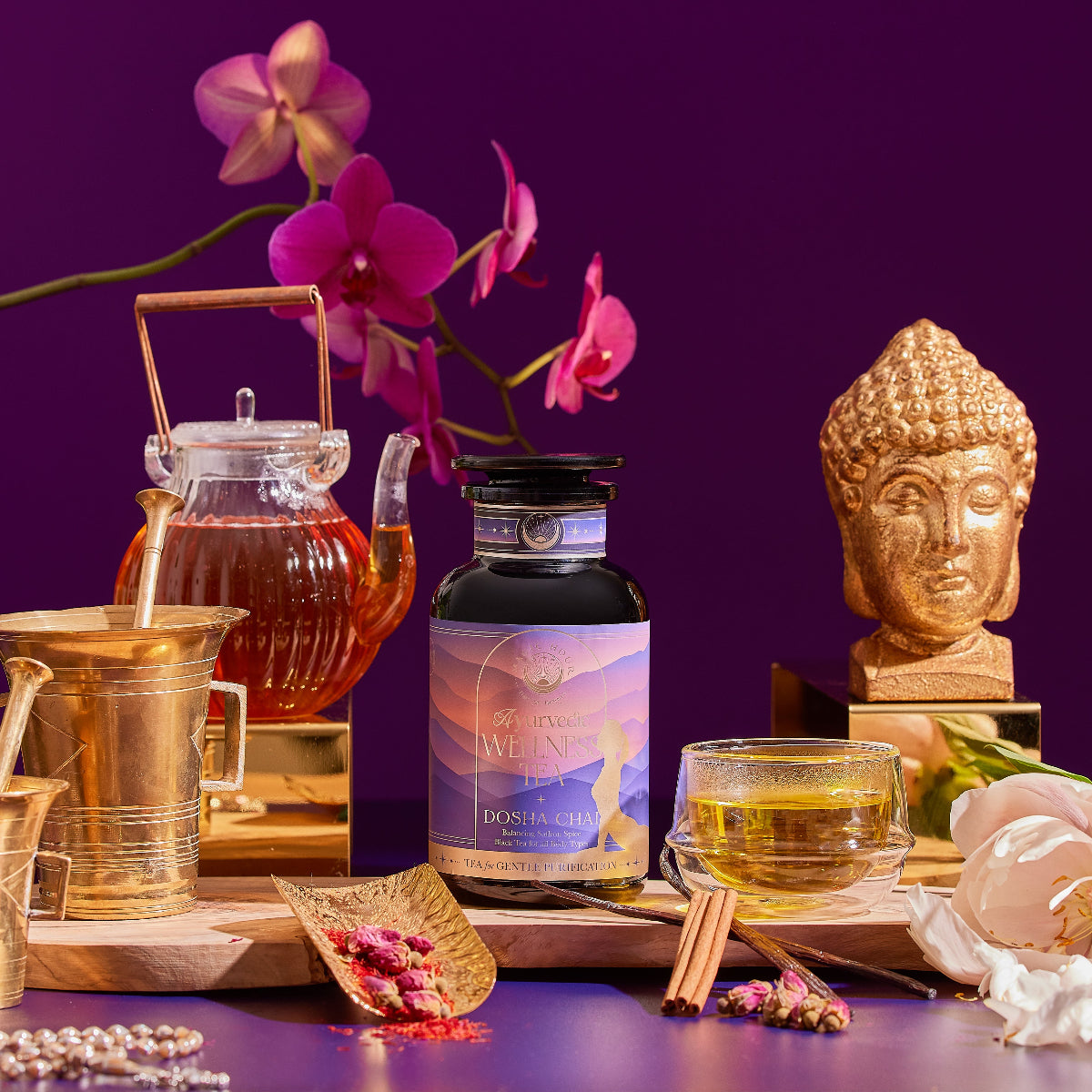 A Dosha Herbal bottle is displayed on a wooden tray with a glass teapot, tea cup, cinnamon sticks, flowers, a gold Buddha statue, and a gold mortar and pestle, set against a purple background with pink orchids.