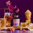 A Dosha Herbal bottle is displayed on a wooden tray with a glass teapot, tea cup, cinnamon sticks, flowers, a gold Buddha statue, and a gold mortar and pestle, set against a purple background with pink orchids.
