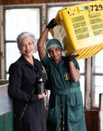 Two women in a well-lit room: one in a black jacket and hair net, holding a water bottle and smiling, the other in green overalls and hair covering, carrying a large yellow crate on her shoulder. They appear to be at Magic Hour's Tea School 2024-2025 Full Course Bundle: Unlock the Secrets of Tea Mastery, possibly participating in tea courses under the supervision of a Master Tea Blender.