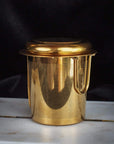 A sophisticated and polished gold metal container from the Magic Hour Starter Kit by Magic Hour is elegantly placed on a white surface against a dark background. The container is adorned with small perforations and features a handle on one side, indicating its potential use for brewing or straining in an elegant tea ceremony.