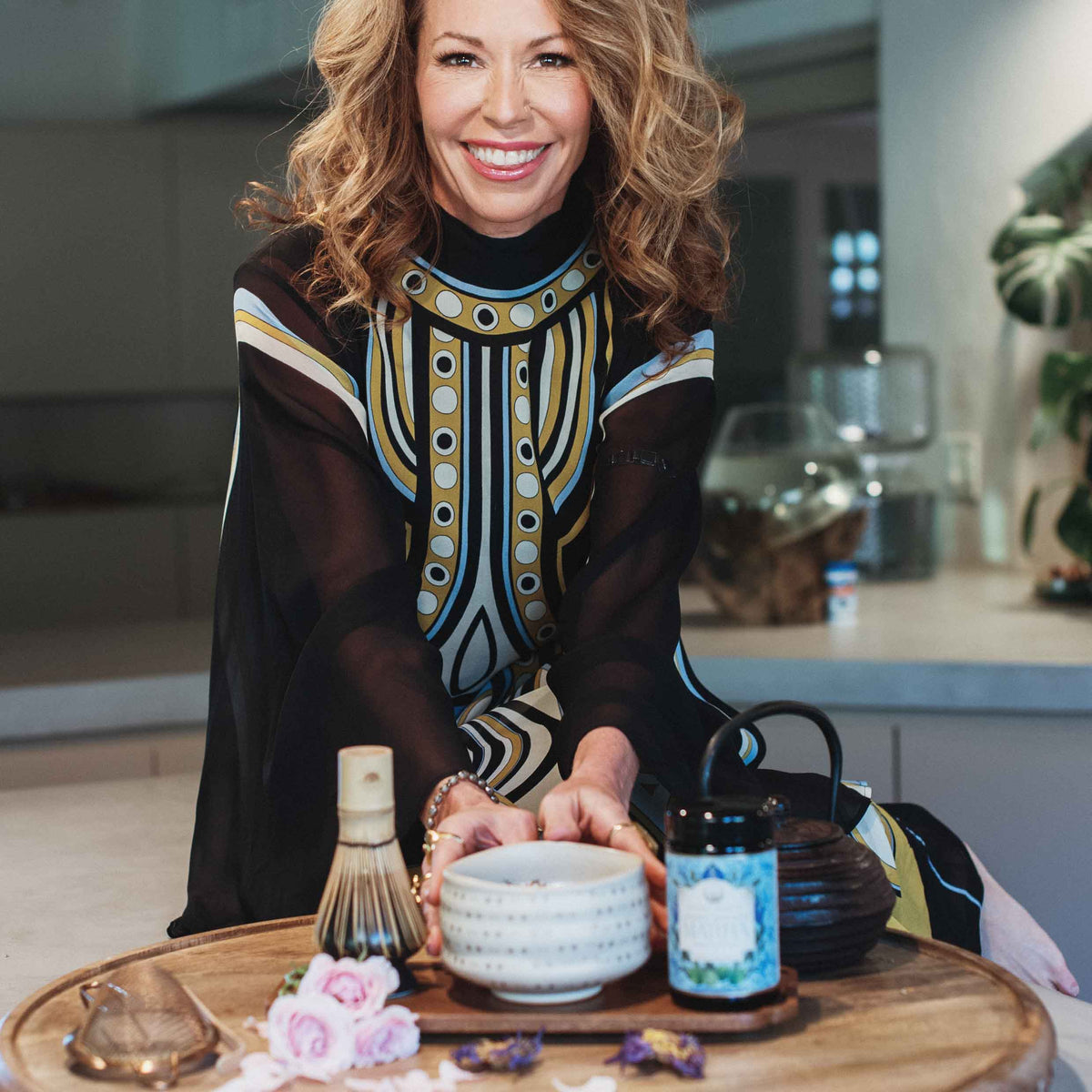 A smiling woman with wavy hair sits at a table with a tea set, featuring the Ceremonial Matcha Bowl—handmade with 14k gold detailing—alongside a jar labeled “cultivate,” a matcha whisk, dried flowers, and decorative items arranged on a wooden tray in a modern kitchen. The scene highlights traditional artisan craftsmanship and warm contemporary style.