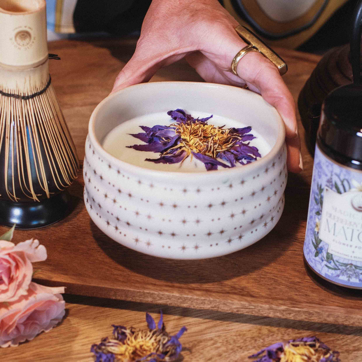 A hand holds the Ceremonial Matcha Bowl, its handmade ceramic surface adorned with delicate star patterns and filled with a creamy matcha drink topped with purple and yellow dried flowers. Nearby, a bamboo whisk, a matcha tin, and pink blossoms rest on a wooden table, capturing the essence of traditional artisan craftsmanship.