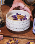A hand holds the Ceremonial Matcha Bowl, its handmade ceramic surface adorned with delicate star patterns and filled with a creamy matcha drink topped with purple and yellow dried flowers. Nearby, a bamboo whisk, a matcha tin, and pink blossoms rest on a wooden table, capturing the essence of traditional artisan craftsmanship.
