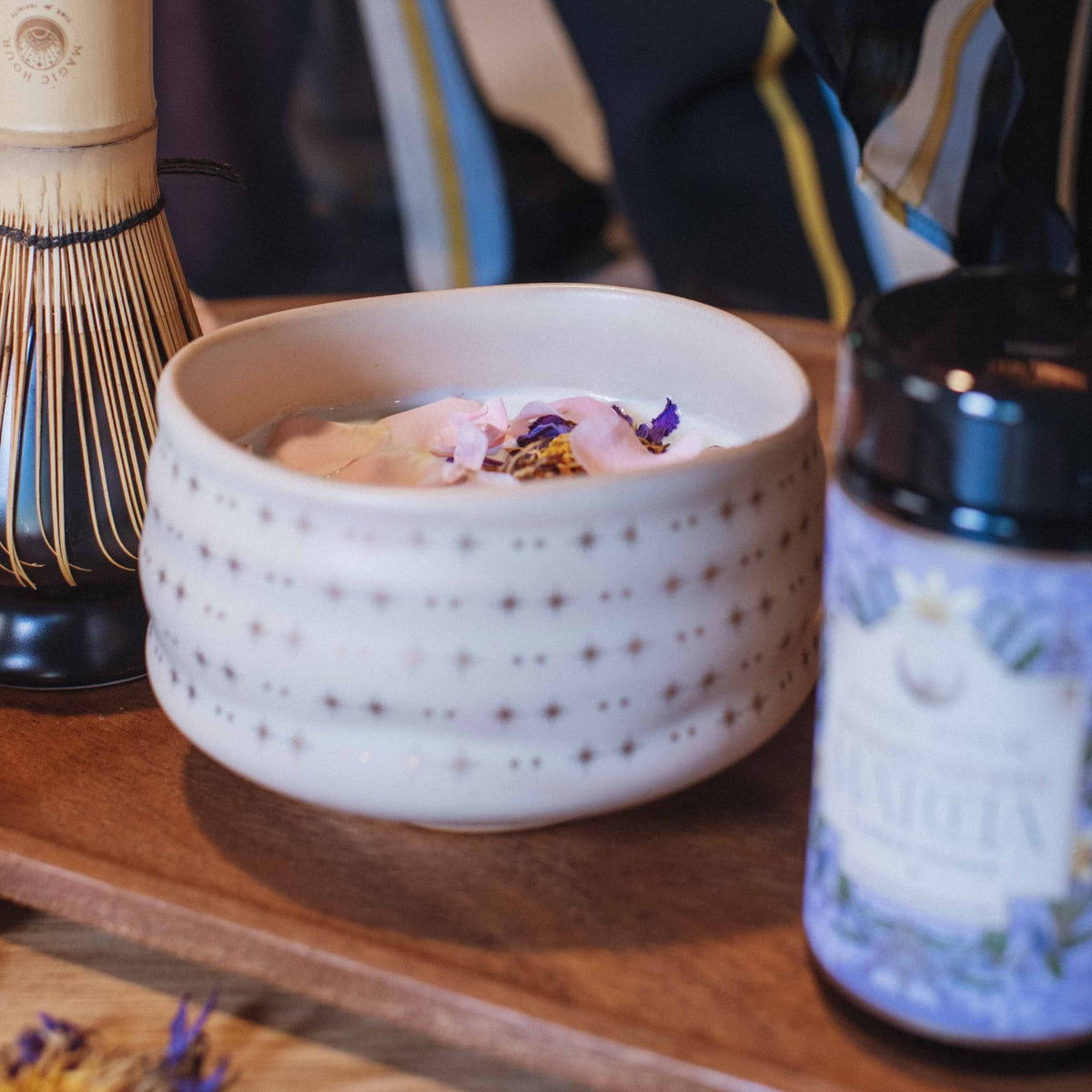 The Ceremonial Matcha Bowl, adorned with delicate star patterns, holds a frothy matcha drink crowned with flower petals. Set on a wooden tray alongside a jar and bamboo whisk, it beautifully highlights traditional artisan craftsmanship.