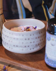 The Ceremonial Matcha Bowl, adorned with delicate star patterns, holds a frothy matcha drink crowned with flower petals. Set on a wooden tray alongside a jar and bamboo whisk, it beautifully highlights traditional artisan craftsmanship.