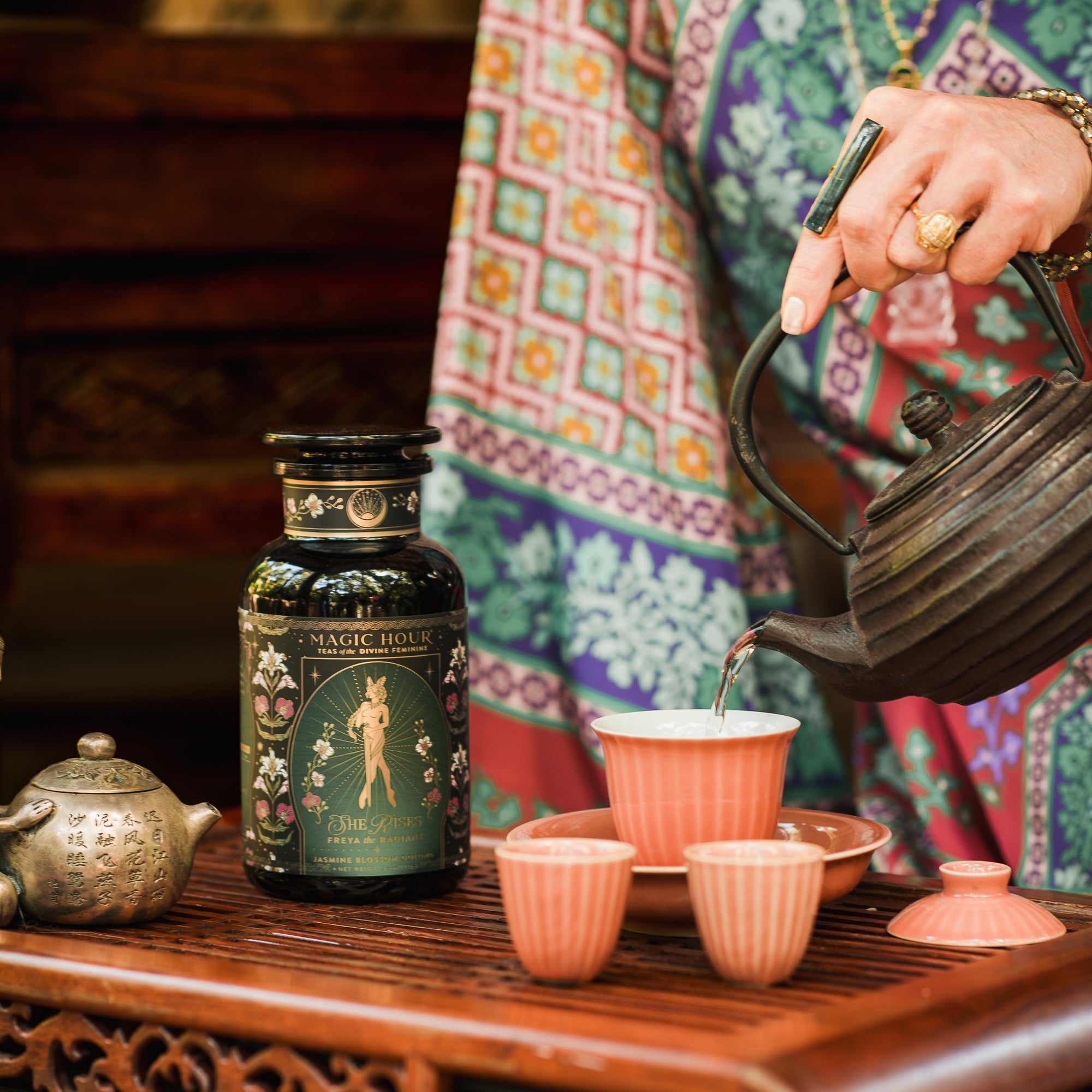 A person in colorful patterned clothing pours hot water into a cup beside a "Freya The Radiant: Jasmine Bai Hao Oolong with Bergamot & Berry" tea jar, with more cups and an ornate teapot on a wooden table.