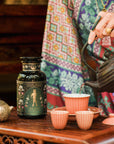 A person in colorful patterned clothing pours hot water into a cup beside a "Freya The Radiant: Jasmine Bai Hao Oolong with Bergamot & Berry" tea jar, with more cups and an ornate teapot on a wooden table.