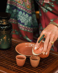 A person in a patterned robe pours tea—perhaps Freya The Radiant: Jasmine Bai Hao Oolong with Bergamot & Berry—from a small teapot into two brown cups on a wooden tray. A dark green tea canister sits nearby. The person wears rings and beaded bracelets.