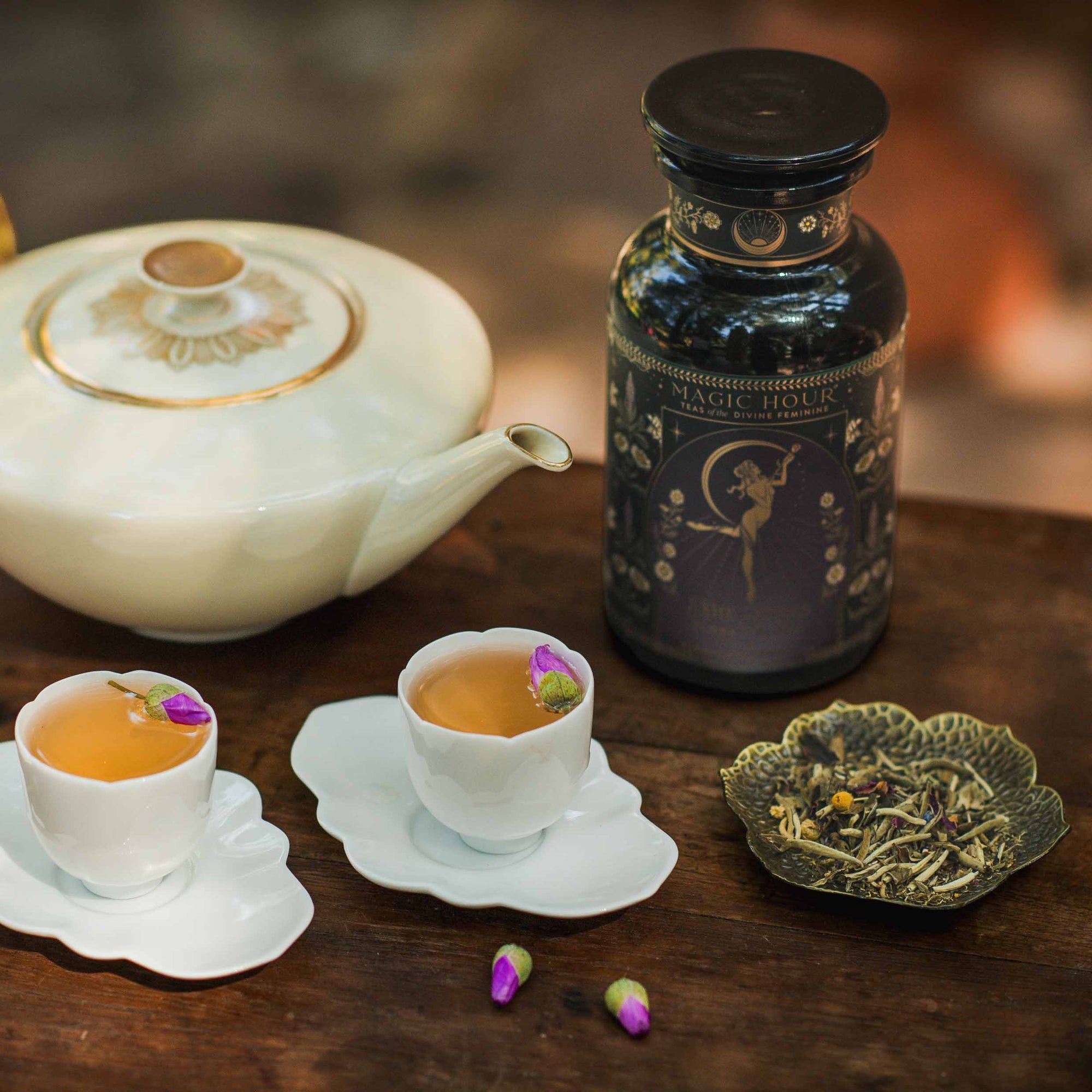 A teapot, two tea cups adorned with rosebuds, a decorative tea canister labeled "Magic Hour," and a plate of loose leaf Flora In Bloom: Peach Moonlight White Tea rest on a wooden table, with scattered rosebuds nearby.