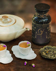 A teapot, two tea cups adorned with rosebuds, a decorative tea canister labeled "Magic Hour," and a plate of loose leaf Flora In Bloom: Peach Moonlight White Tea rest on a wooden table, with scattered rosebuds nearby.