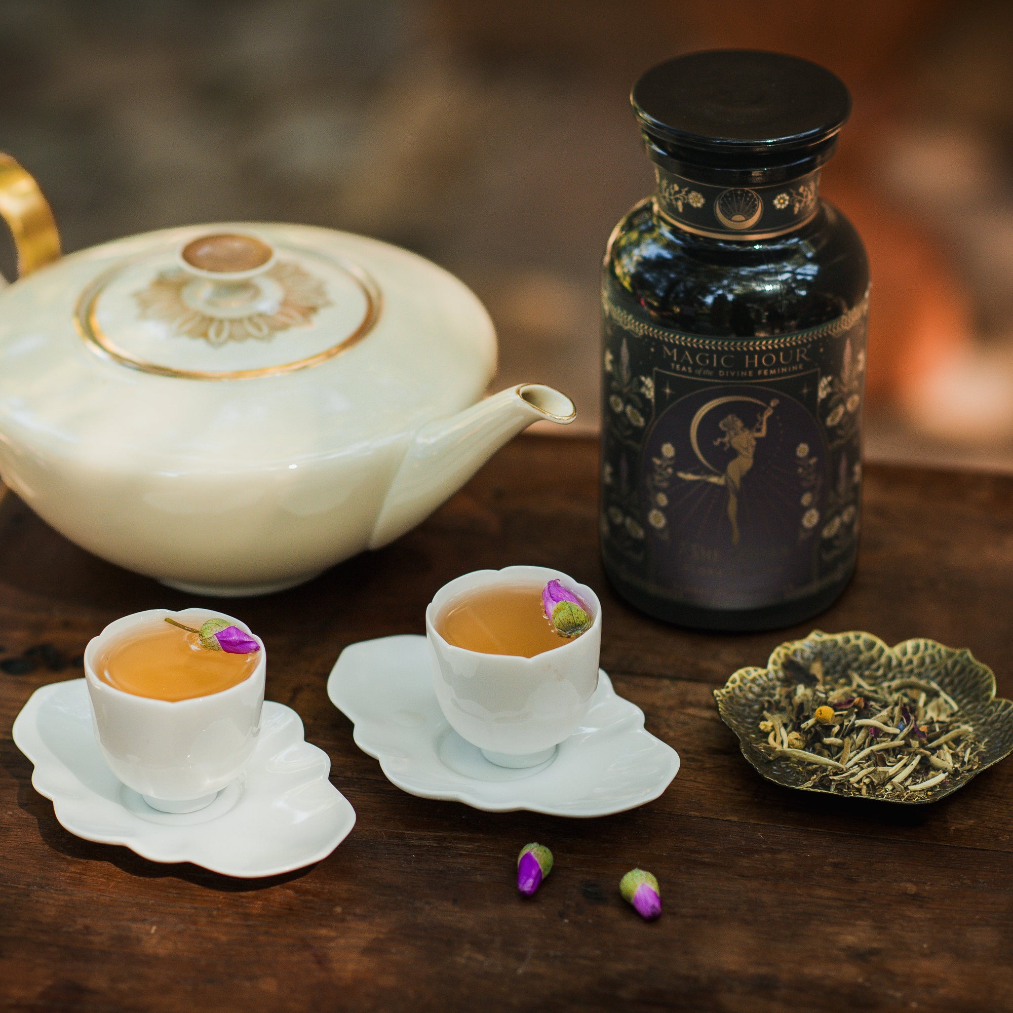 The Flora In Bloom: Peach Moonlight White Tea set features a floral teapot, two rosebud teacups, loose leaf tea on a decorative plate, and a black "Magic Hour" jar, all arranged on a wooden surface.