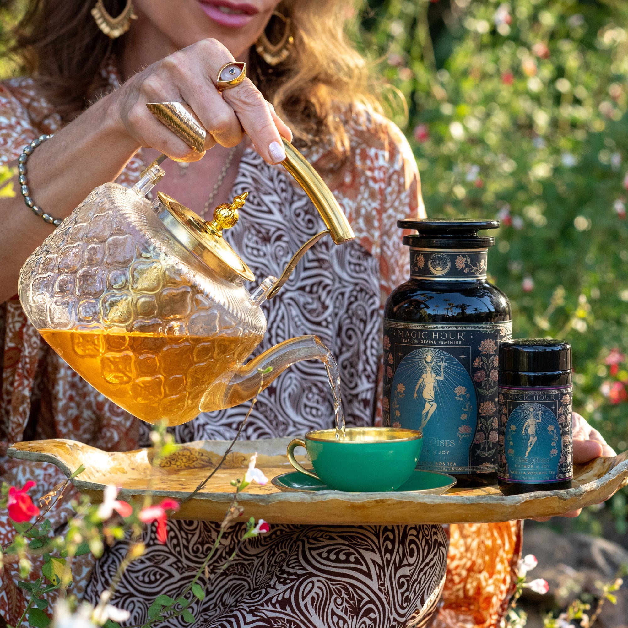 Outdoors among greenery and flowers, someone in patterned clothing pours Hathor of Joy: Honey Nectar Herbal Tea from a glass teapot into a green teacup on a tray, with two ornate "Magic Hour" containers beside the cup.