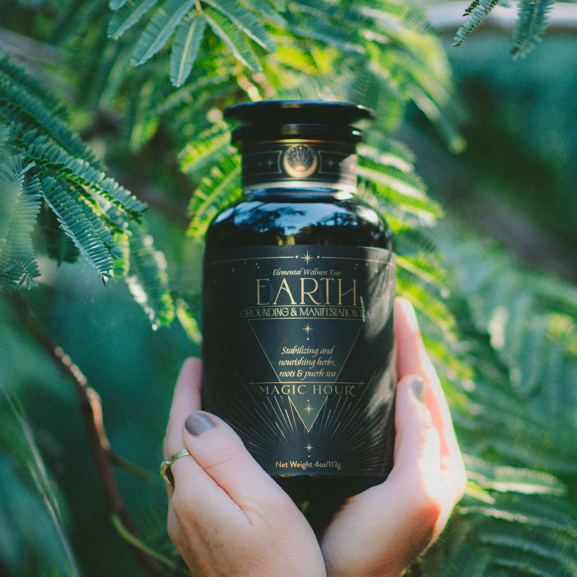 A person with painted nails and rings holds a black jar labeled “EARTH Stabilizing & Nourishing Herbs, Roots, and Puerh Tea for Digestion & Grounding” by Magic Hour—an earthy blend crafted to support grounding and digestion. Green leaves and sunlight can be seen in the background.