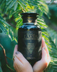 A person with painted nails and rings holds a black jar labeled “EARTH Stabilizing & Nourishing Herbs, Roots, and Puerh Tea for Digestion & Grounding” by Magic Hour—an earthy blend crafted to support grounding and digestion. Green leaves and sunlight can be seen in the background.